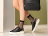 Person wearing black sneakers with white soles and sheer black socks, standing against a green wall.