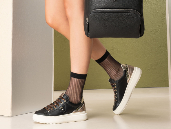 Person wearing black sneakers with white soles and sheer black socks, standing against a green wall.